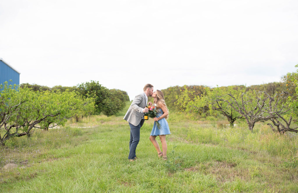 Hancock Orange Groves Engagement Session Couple holding flowers and champagne kissing celebrating in a green orange field