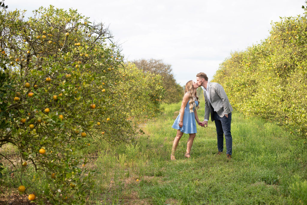 Hancock Orange Groves Engagement Session Couple leaning towards each other kissing in a orange farm wearing a short light blue dress and a grey tailored suit 