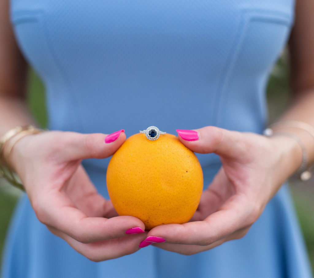 Hancock Orange Groves Engagement Session Woman holding Blue Wedding Engagement Ring Silver on top of an orange in a blue dress