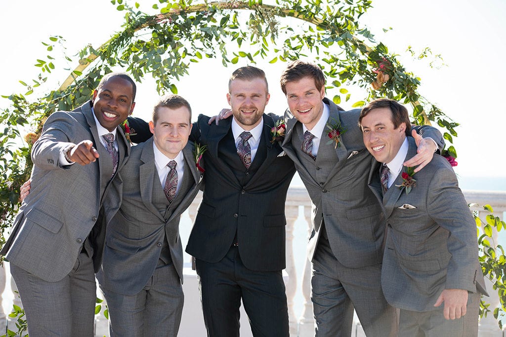 Hyatt Regency Clearwater Beach Wedding Groom in black tuxedo from Jos A. Banks and groomsmen in grey tuxedos smiling looking at the camera on the sky terrace with the water in the background and the floral moon gate arch