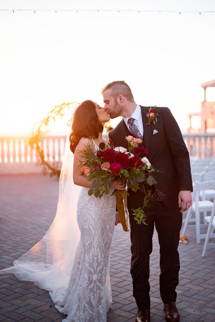 Hyatt Regency Clearwater Beach Wedding Bride wearing Galia Lahav sicilian dress GALA collection holding big red and pink color flowers bouquet with yellow details kissing groom in black tuxedo in the sky terrace by the water with floral moon gate arch