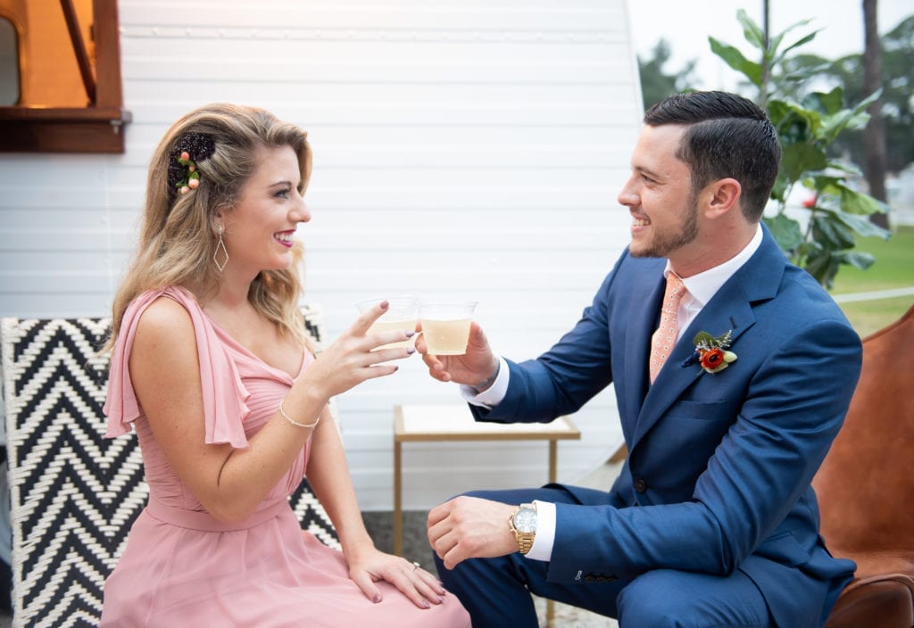what to wear to your engagement session woman in pink dress sitting down next to man in navy blazer sitting down holding a drink doing cheers and smiling