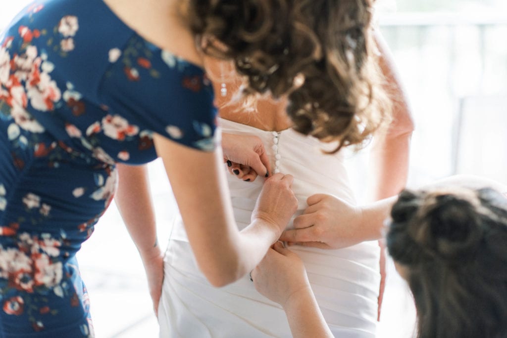 Bridesmaids helping bride put her dress on and get ready by buttoning her dress on