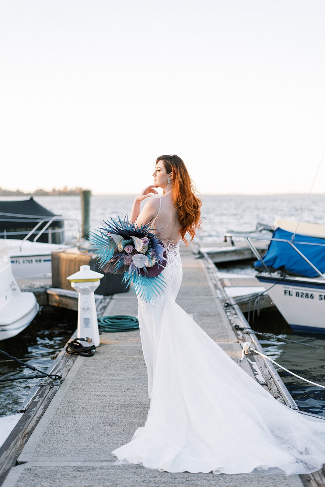 Top Reasons To Hire A Wedding Planner with The little mermaid inspired wedding at mission inn resort bride by the boat dock posing with her purple and blue wedding bouquet looking off in the distance and her back is to the camera