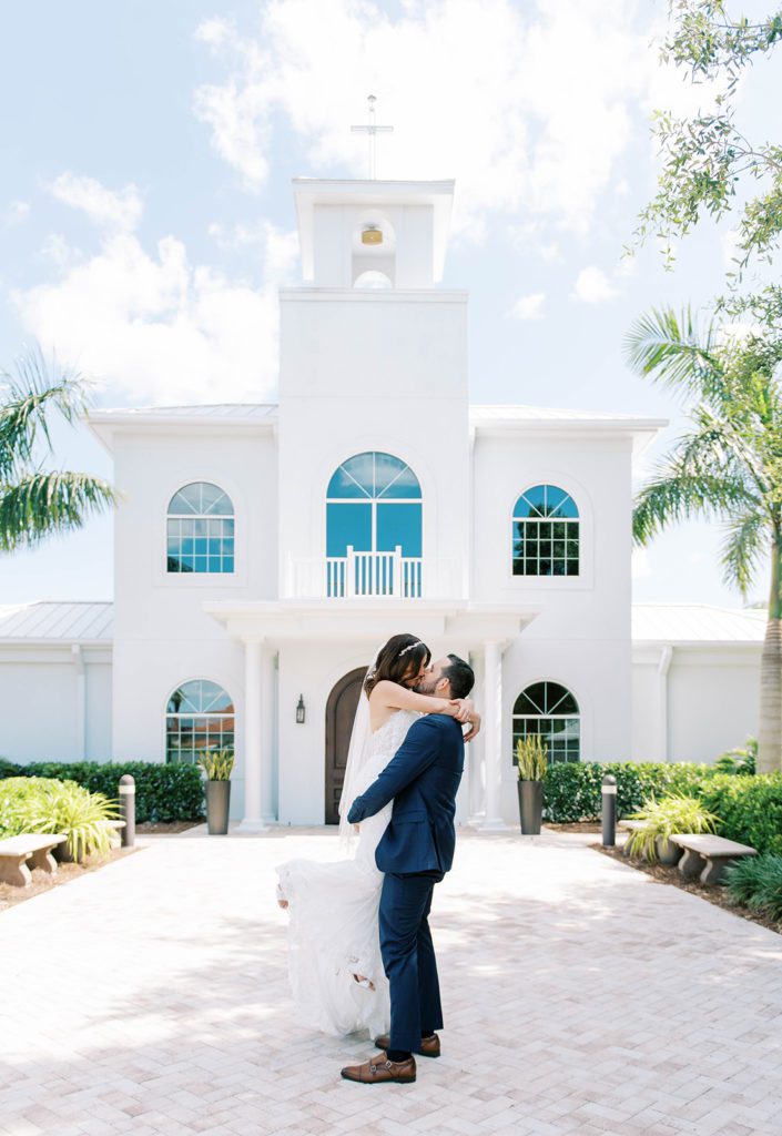 bride and groom standing on the lawn of a church building with groom picking up bride and kissing her has she wraps her arms around his neck wedding photography timeline
