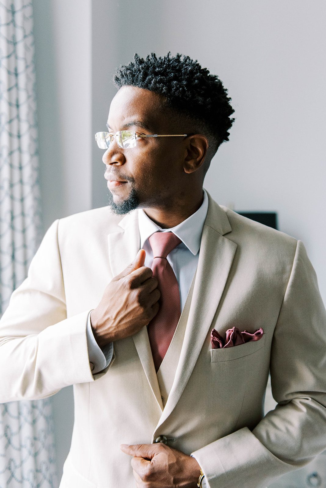 man in a tan suit with a light pick tie adjusts his suit and tie as he looks out of a window as he gets ready for his wedding day in Tampa Florida