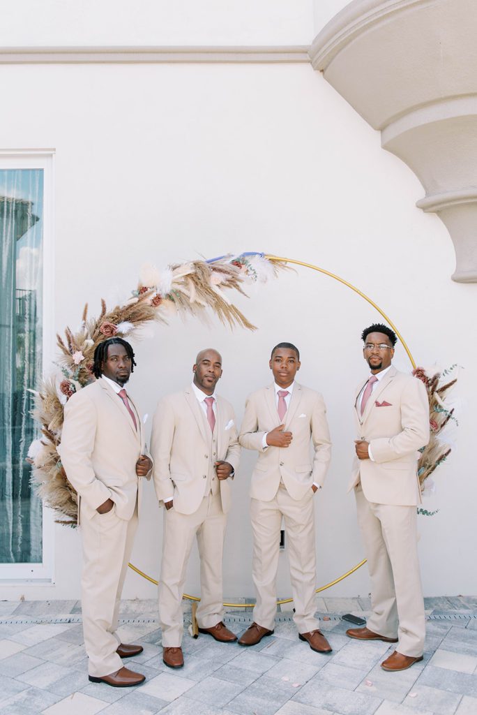 groomsmen standing under a wedding arch in cream suits with blush pink ties and pocket squares in Tampa Florida wedding