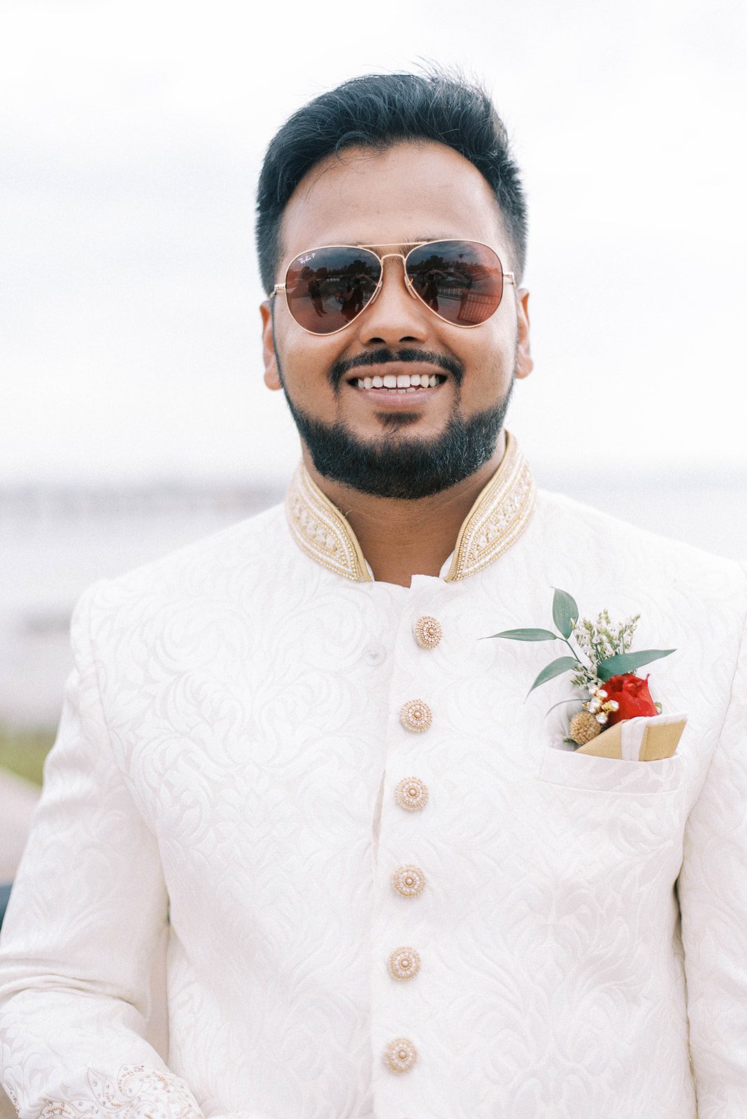 Indian groom smiling while wearing aviators and his traditional Indian wedding outfit that is white with beaded details and a red carnation in his coat pocket