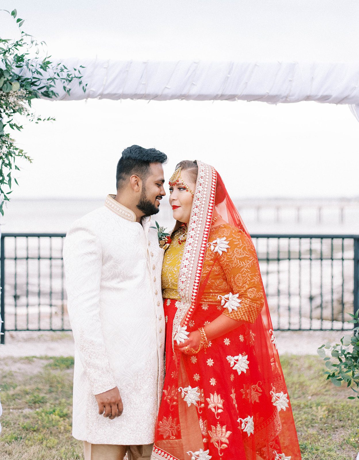 Indian bride and groom standing under a wedding arc hnext to the ocean as they look into each others eyes and smile while wearing their tradtional Indian wedding apparel