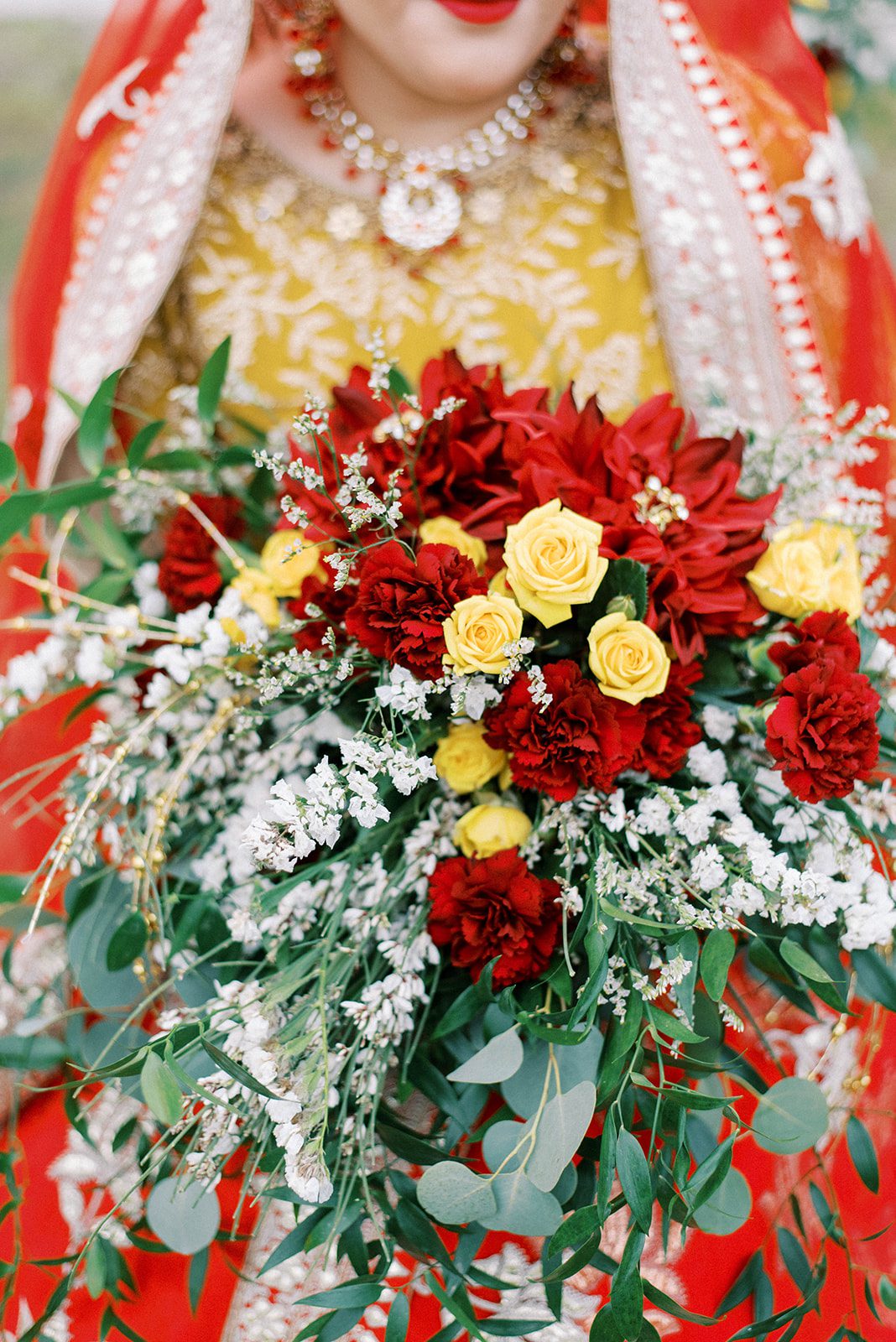 Indian Bride in red and yellow accents holding a red rose bouquet with lush greenery and yellow and white accent flowers