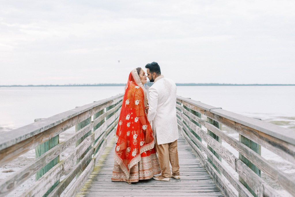indian bride and groom in traditional Indian wedding apparel walking on a dock into the ocean as teh lean into a kiss 