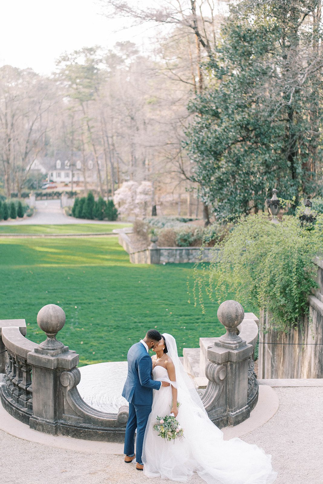 bride and groom standing on a lower balcony of a palace styled mansion that looks over a green garden as they kiss each other of their destination wedding day Swan House Atlanta Wedding
