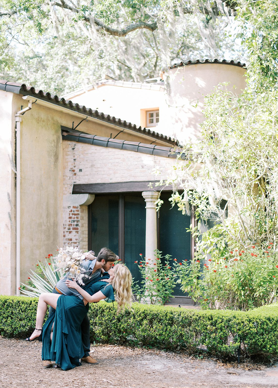 man and woman in Bok Tower Gardens for their engagement session with Tampa wedding photographer, they are infront of a smaller structure and garden landscape with the man dipping the woman as her emerald green gown's slit opens to reveal her leg