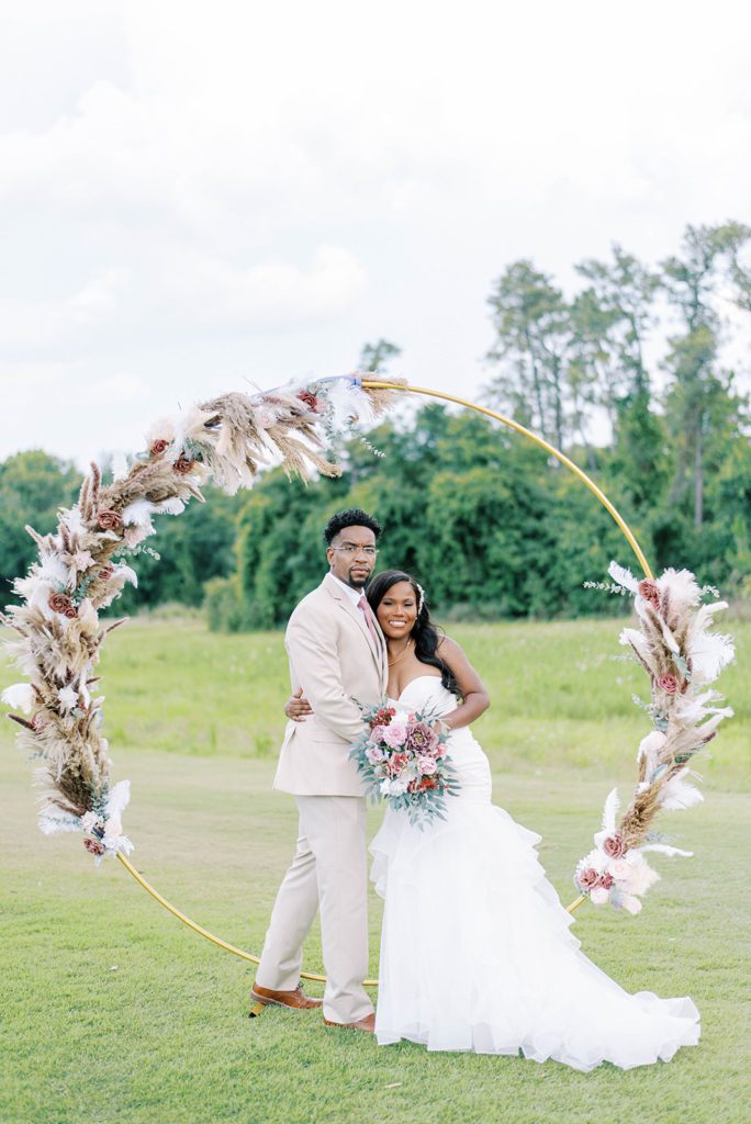 bride in a sweetheart neckline drop waist gown with her groom in his cream suit under a circle wedding arch that had pompous grass and blush roses to decorate the arch