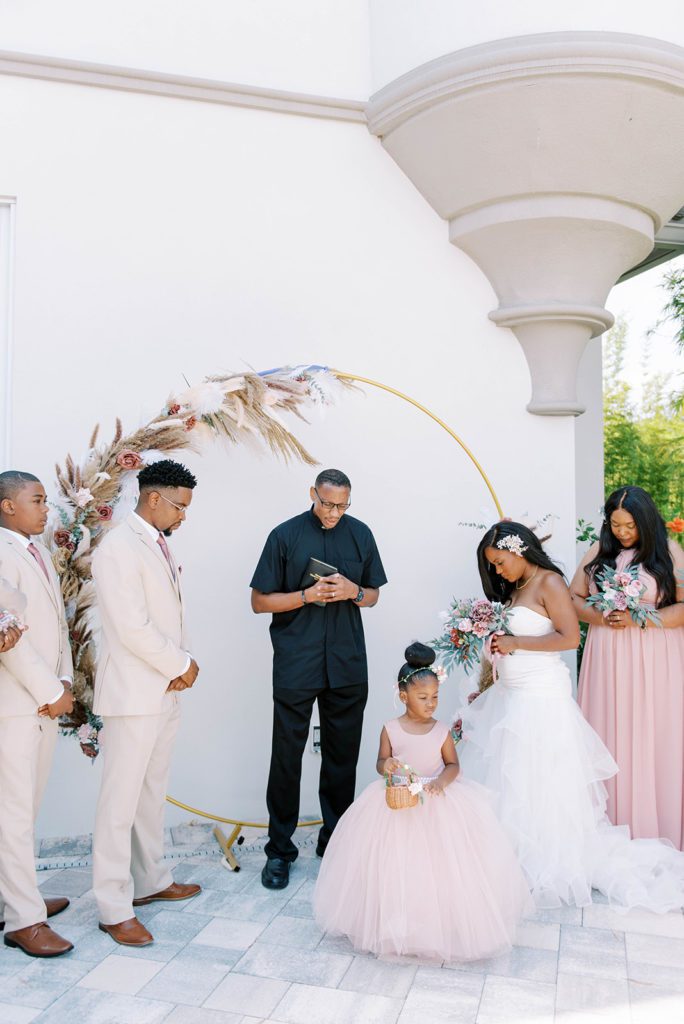 Airbnb wedding ceremony bridesmaids in dusty rose groomsmen in cream tuxedos bride in white dress next to officiant praying during the ceremony
