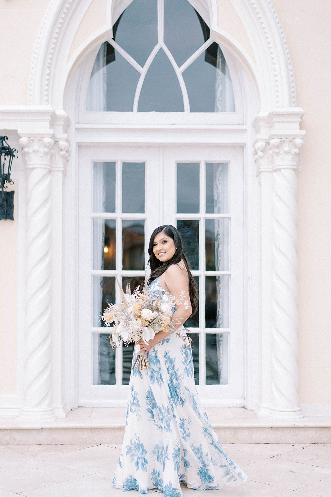 engagement session ay the Mirasol with woman standing in a floor length blue and white gown with floral details and holding a neutral tones bouquet and smiling over her shoulder