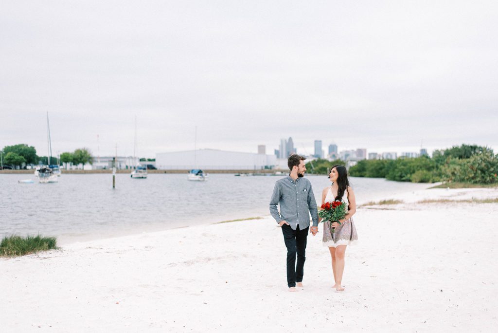Davis Island engagemtn session on the beach with man in blue shirt and woman in neutral summer dress holding hands and walking together on the white sand with sail boats behind them