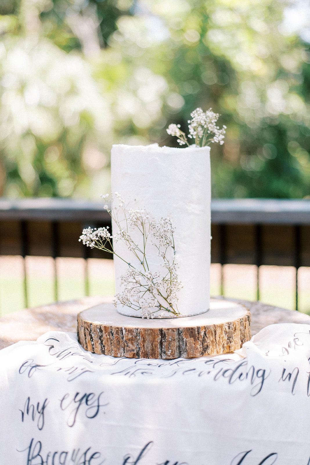 stunning white minimalist wedding cake with small florals on the side on the cake while it sits on a wooden round cake stand and a tapestry underneath