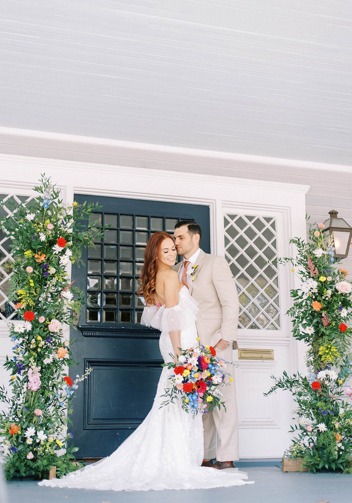 retro inspired wedding at the Orlo wedding venue in Tampa Florida with bride in a lace gown and groom in a cream suit embracing and the groom kissing the bride's cheek on a front porch