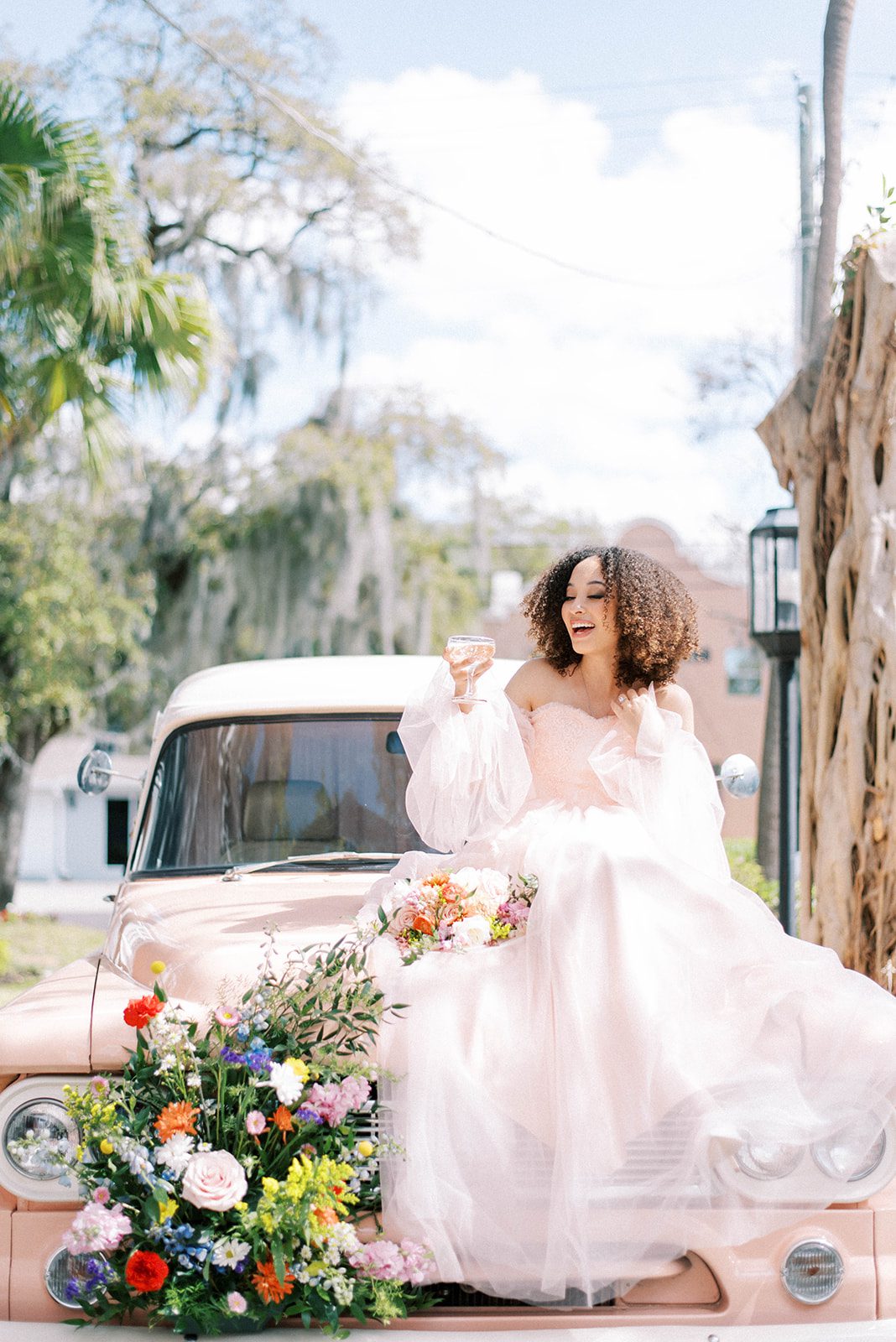 bride in a pastel pink tulle wedding dress sitting on a vintage pink car while sipping champagne out of a fancy cup with a bright and full bouquet of florals sitting at the brides feet 2022 Wedding Trends
