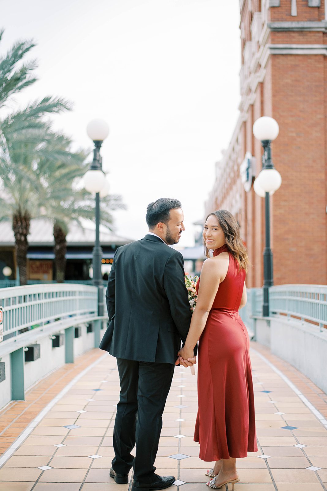engaged couple walking on a bridge in Ybor City while holding hands and looking over their shoulders back at the camera, the woman is in a red mid length gown and the man is in a black suit with no tie, and old historic building surround them