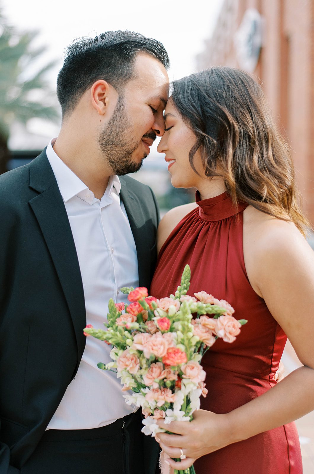 man and woman who are engaged embrace with one arm and the man rests his forehead on the woman's and they both smile as the woman holds a pink rose bouqet with brick buildings behind them