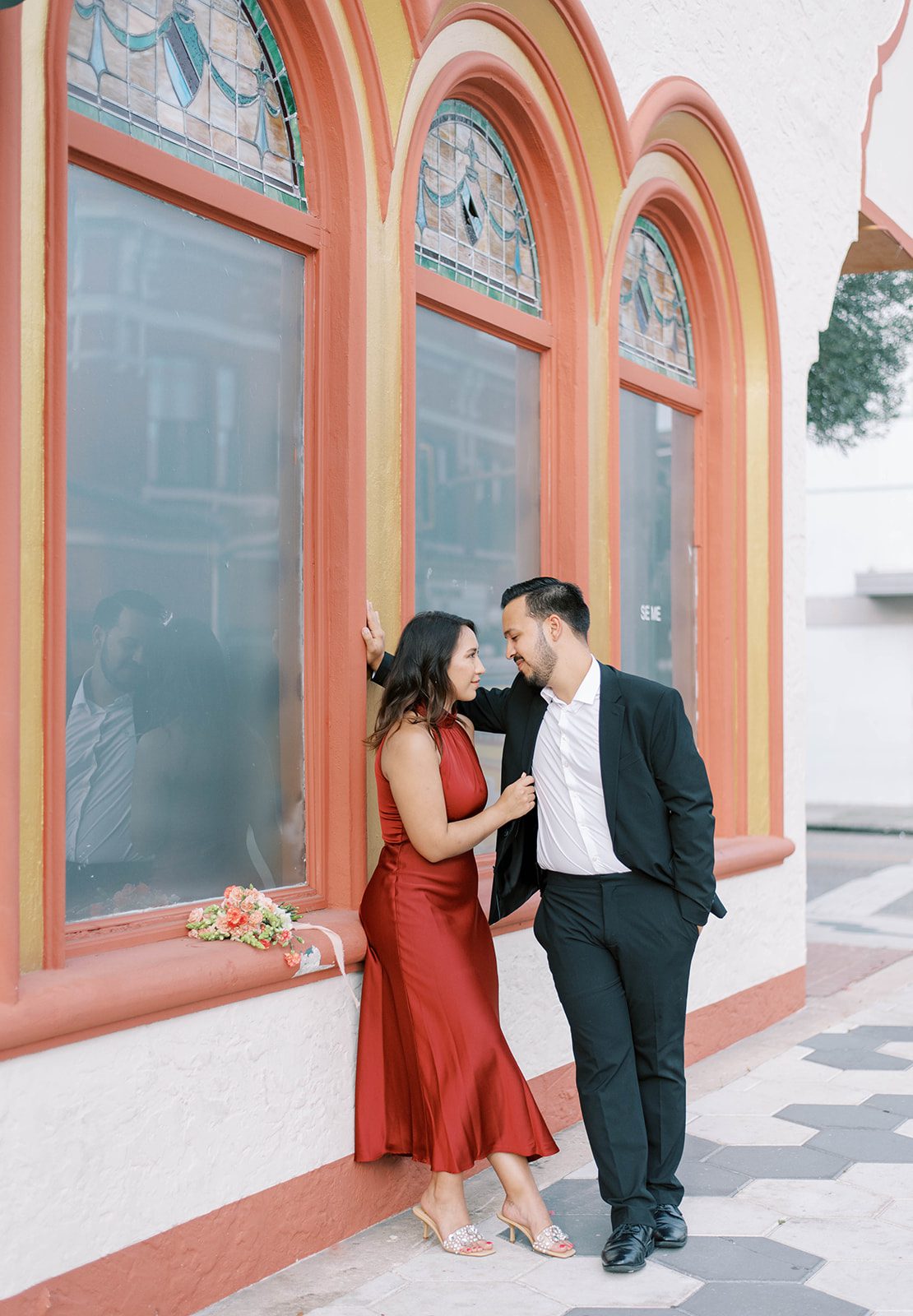 Ybor City engagement session with man in black suit and no tie leaning against a wall with a hand in his pocket over his fiance as her back is against the wall and she is holding his suit coat, with stunning old glass window behind them