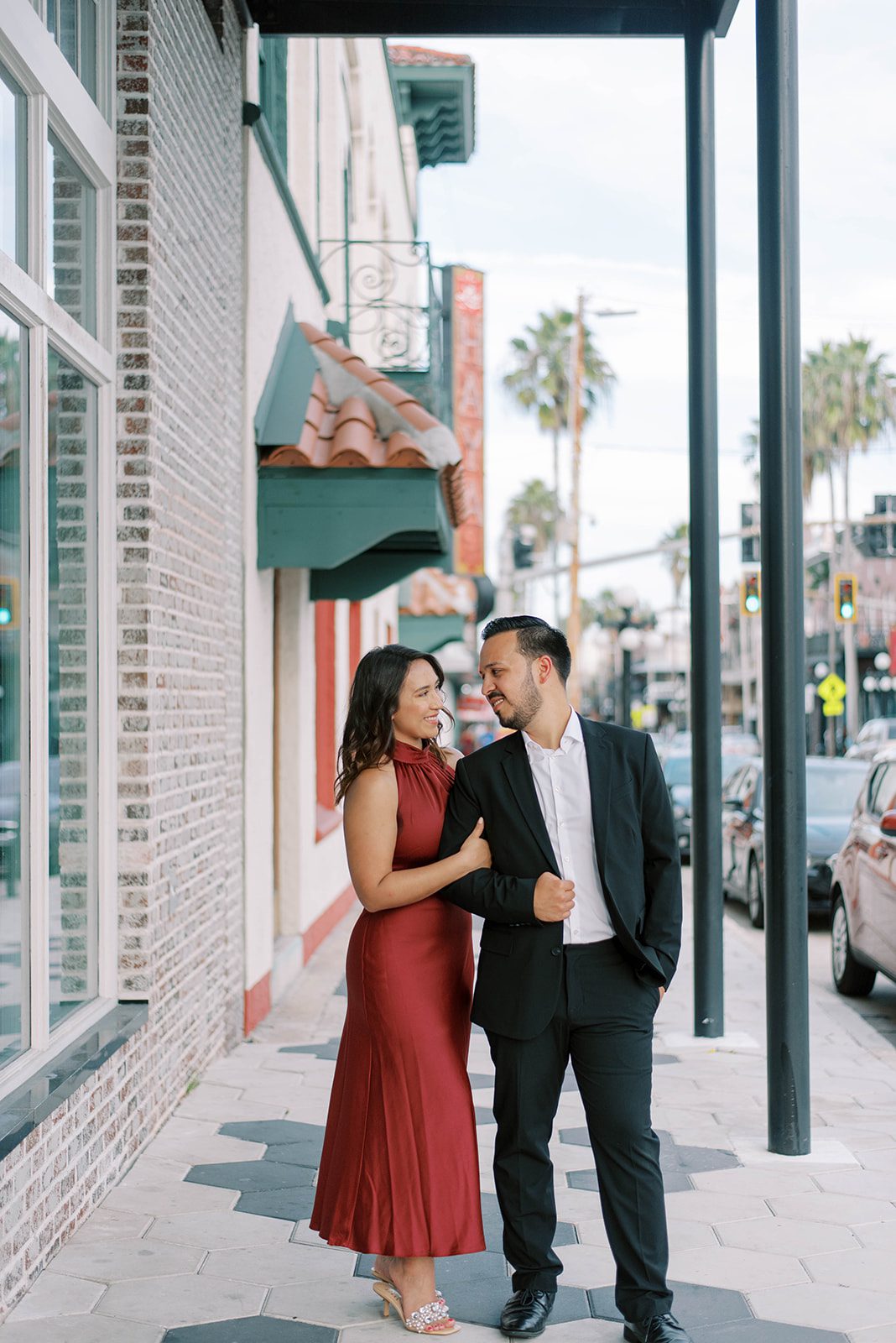 woman and man on the street of Ybor City down town with woman holding mans arm as she stands next to his and they both look at each other romantically, woman in a red dress for her engagement session