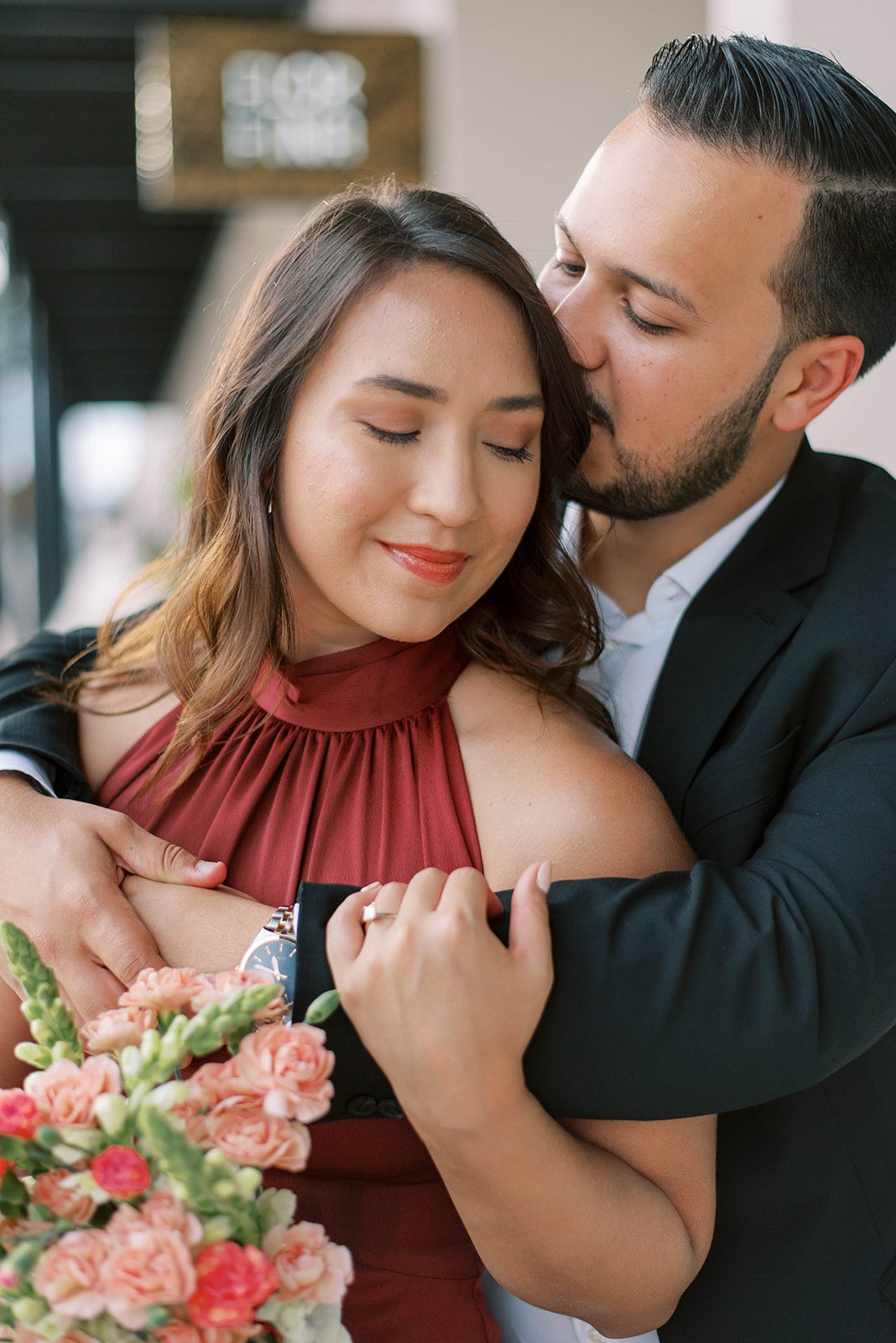 man standing behind his fiance with his arms around her shoulders and kissing her on the head as she holds his arm and smiles while looking down