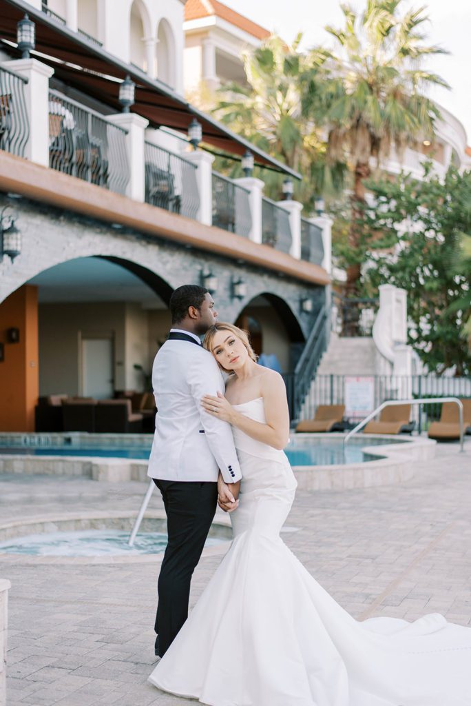 bride holding her grooms arm and looking over his shoulder at the camera as the groom stands facing away from the camera in front of a large Spanish inspired building