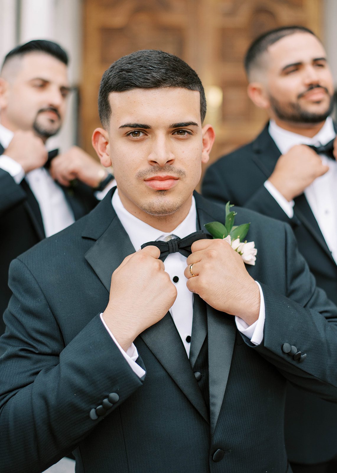 Latino groom fixing his bow tie as he looks directly into the camera with a smolder while standing outside the wedding chapel in Tampa Florida