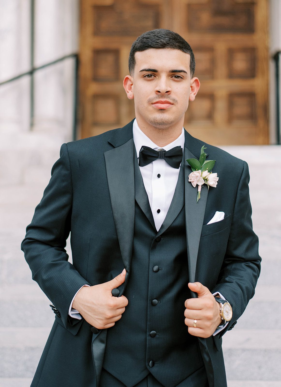 groom holding his black tuxedo and looking into the camera with the large wood doors of a cathedral behind him in Tampa Florida