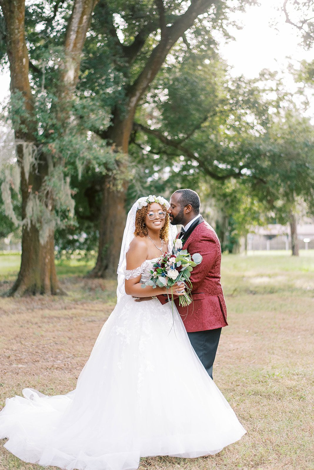 bride in a large fluffy white gown with a stunning red and white bridal bouquet is held by her groom who is wearing a red suit coat as they stand outside of their wedding venue