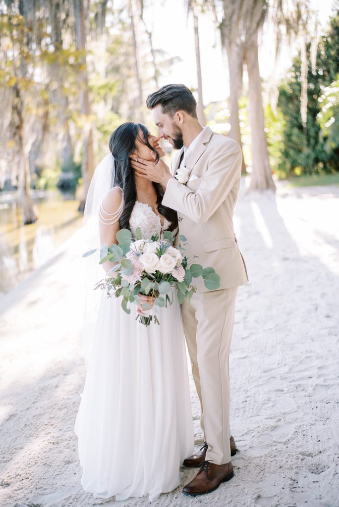 groom in a beige suit holding his bride face as she looks up at him and smiles while holding her wedding bouquet with one hand and him with the other, captured by the best Tampa Bay wedding photographers