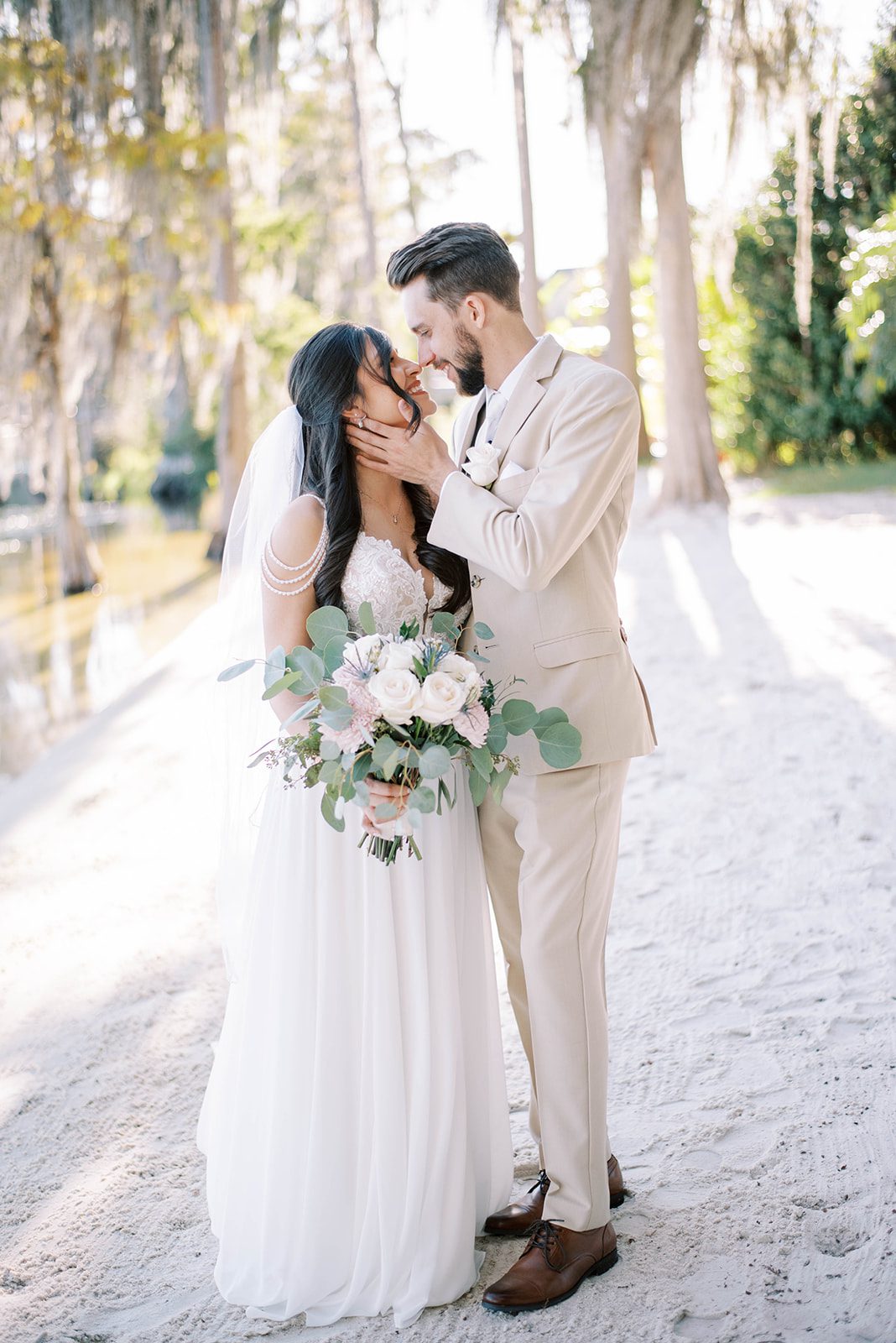 groom in a beige suit holding his bride face as she looks up at him and smiles while holding her wedding bouquet with one hand and him with the other, captured by the best Tampa Bay wedding photographers