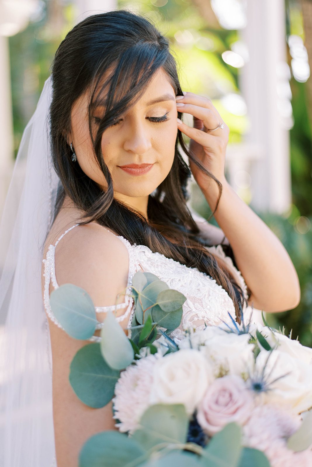stunning dark haired bride holds her wedding bouquet that is full of bright white and light pastel roses while tucking her hair behind her ear and slightly smiling as she looks down over her shoulder, taken by best Tampa wedding photographers