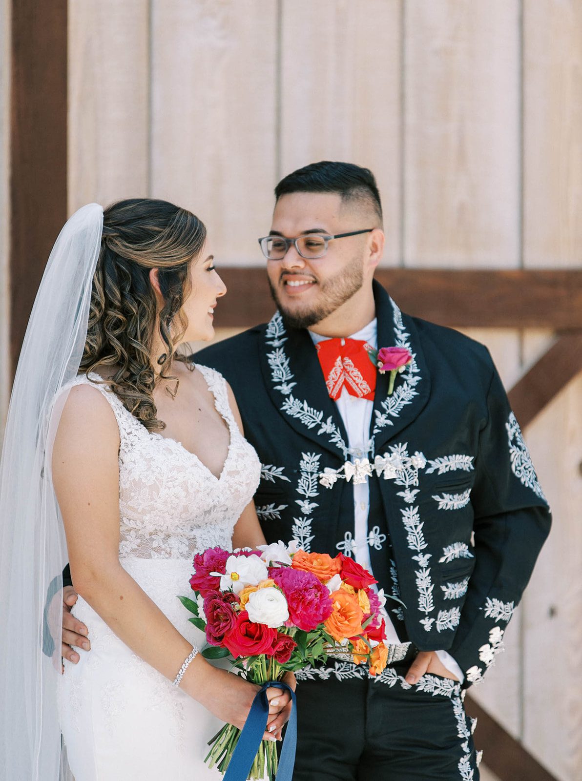 Mexican bride and groom smiling at one another as they stand outside of a barn together.