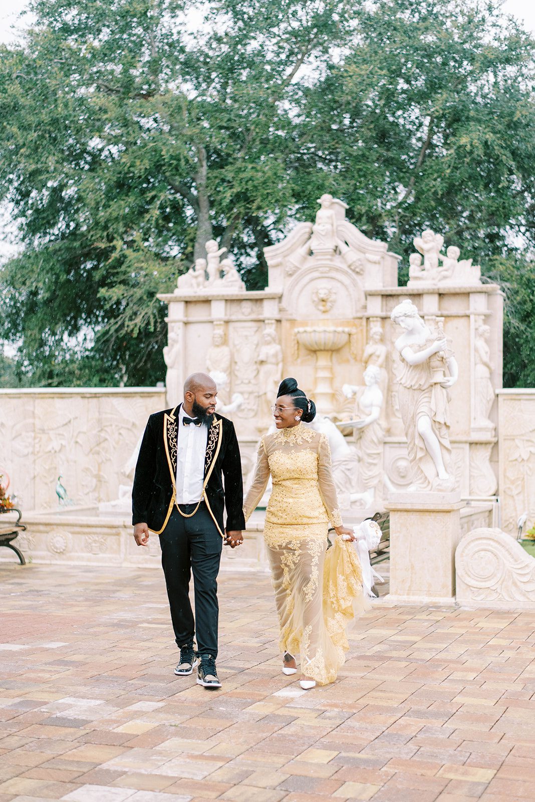 bride in a gold lace wedding dress holding hands with her groom as they walk towards the camera outdoors at their luxury wedding venue
