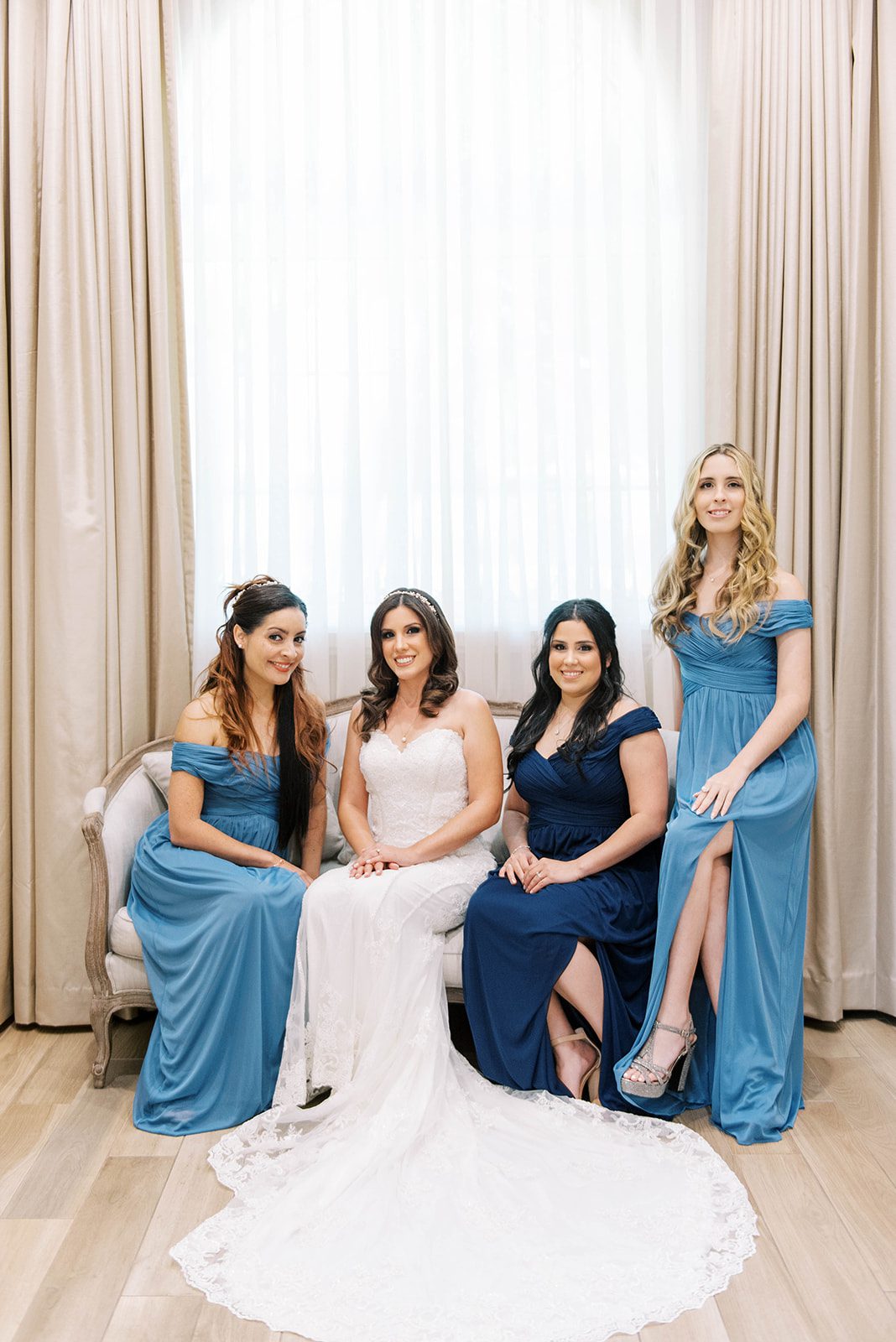 bride sitting on an elegant sofa with her bridesmaids who are in different shades of blue after they have gotten ready and before the wedding ceremony for Tampa bridal photography