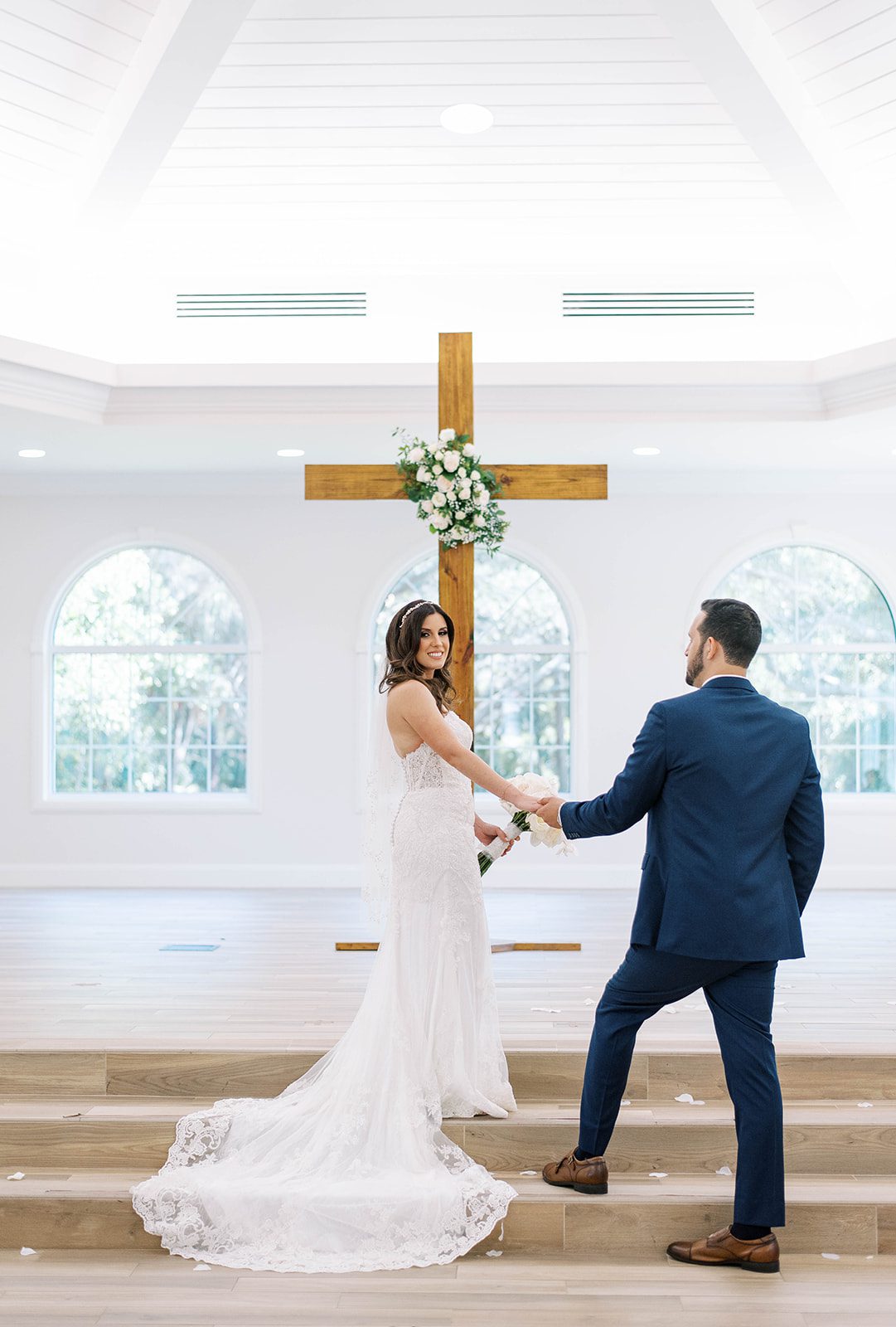 Harborside Chapel wedding in Tampa Florida with bride in a lace wedding dress holding hands with her groom as she walks up the stairs on the chapel's stage and smiles at her groom