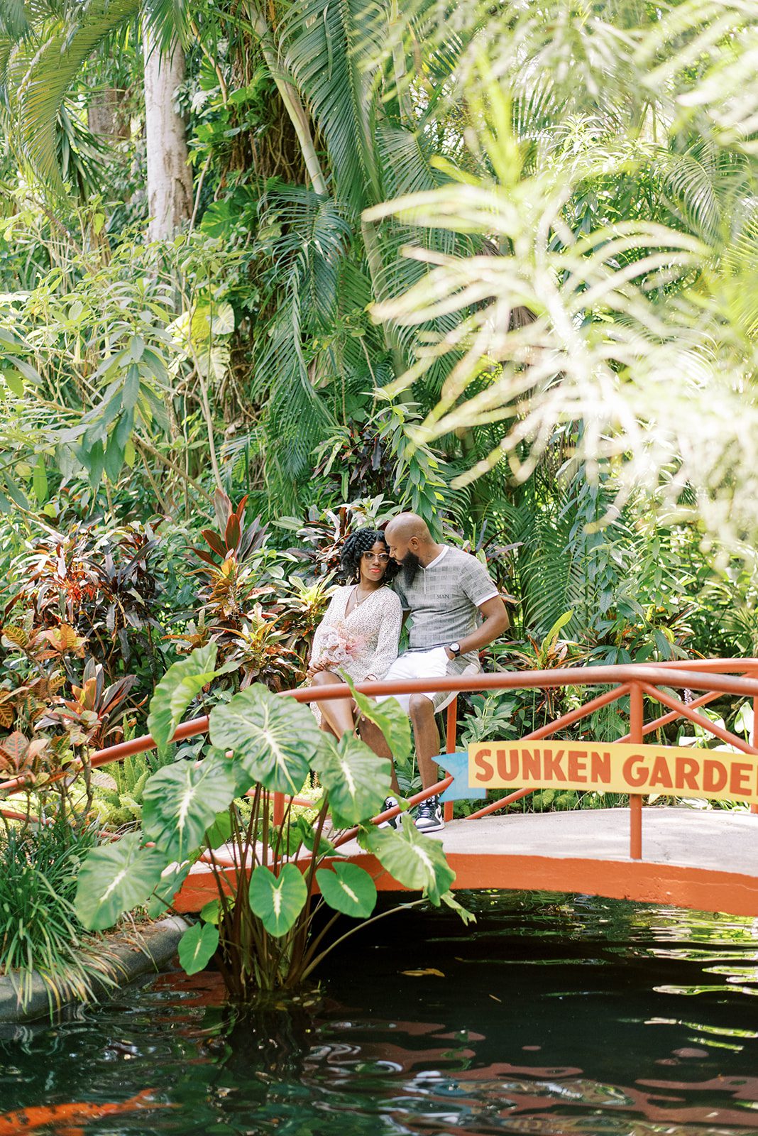engagement photos with man and woman sitting on a bridge together in a lush Florida garden with water flowing under them in a stream