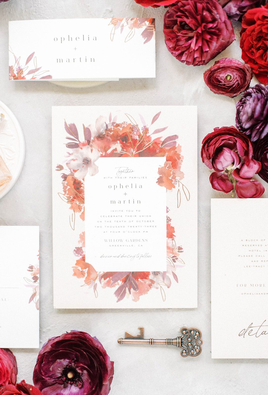 wedding invitation in white and a deep red surrounded by wedding flowers and a key detail