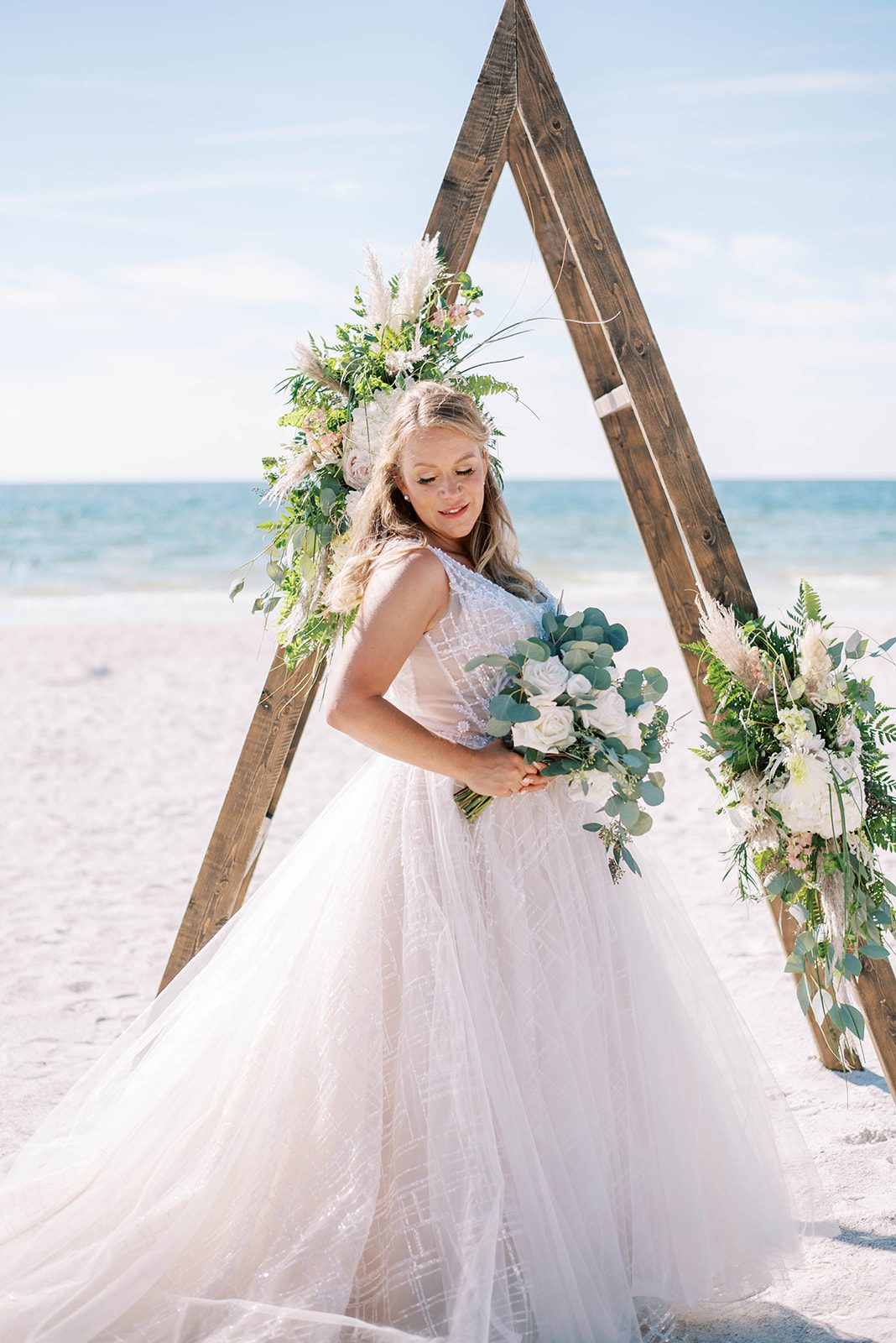 beautiful bride on a beach in Florida posing with her wedding bouquet in front of her triangle wedding arch