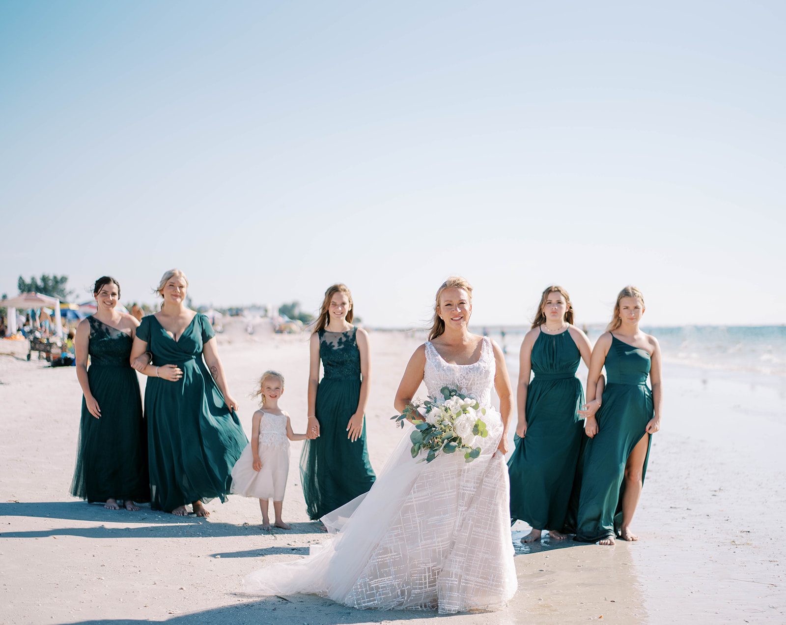 bride walking on the beach with her bridesmaids walking behind her as they wear Juniper green bridesmaids dresses for their St. Petersburg Florida beach wedding