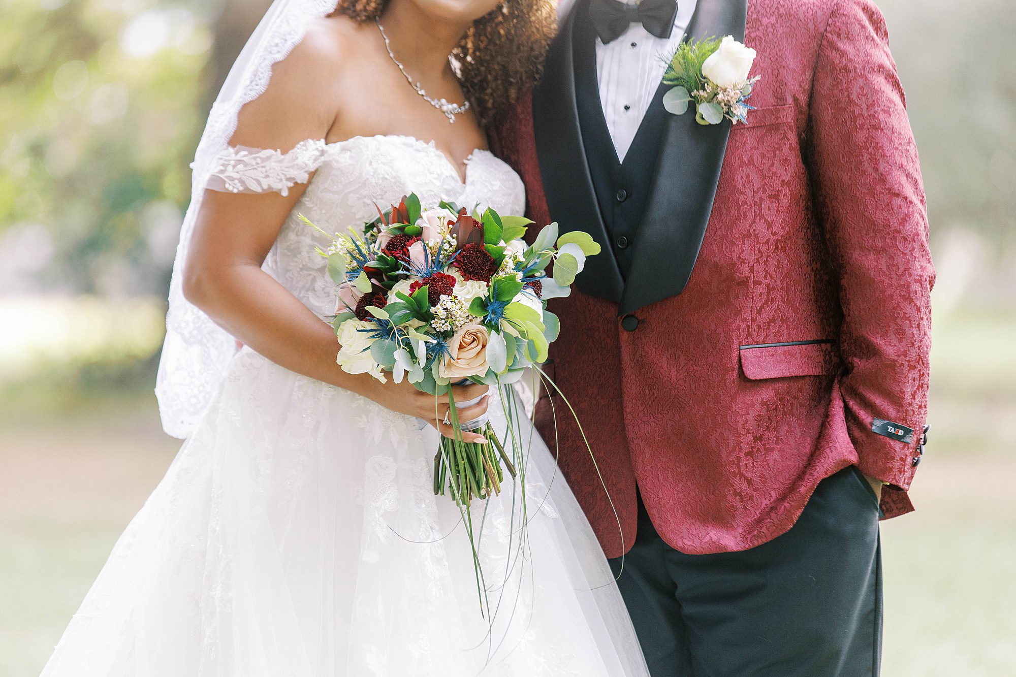 bride hugs groom in red suit jacket holding bouquet of pink and red flowers