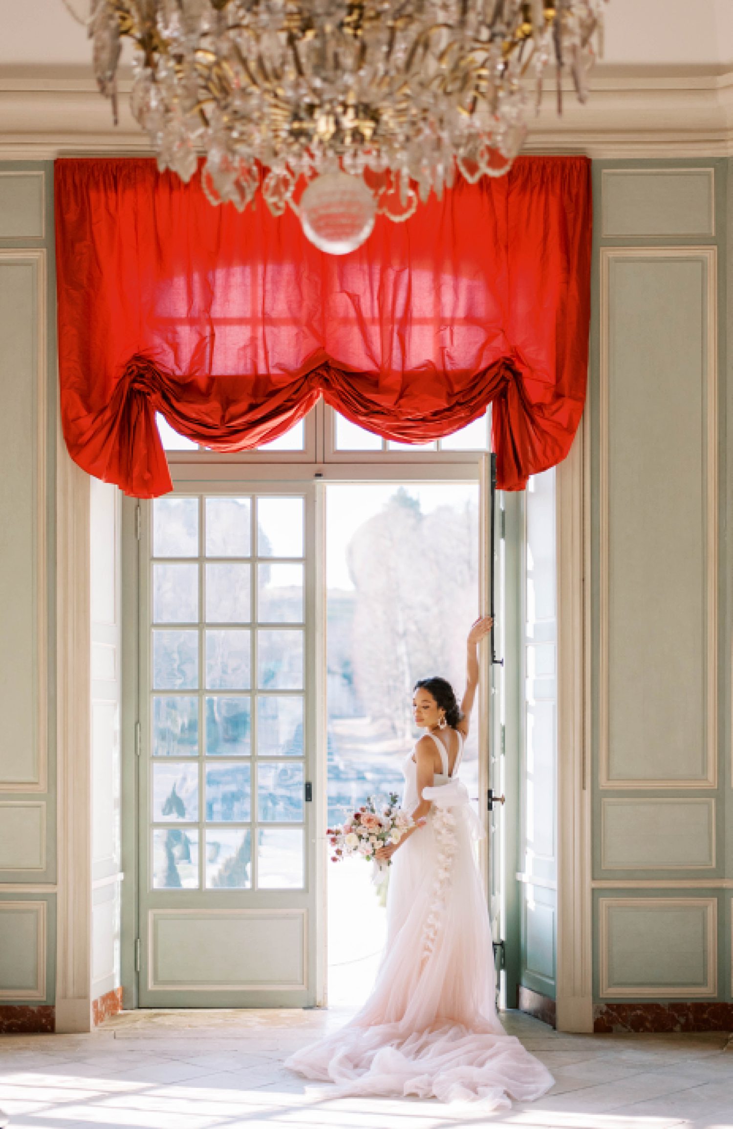 bride stands in doorway under red curtain in blush Marchesa wedding gown