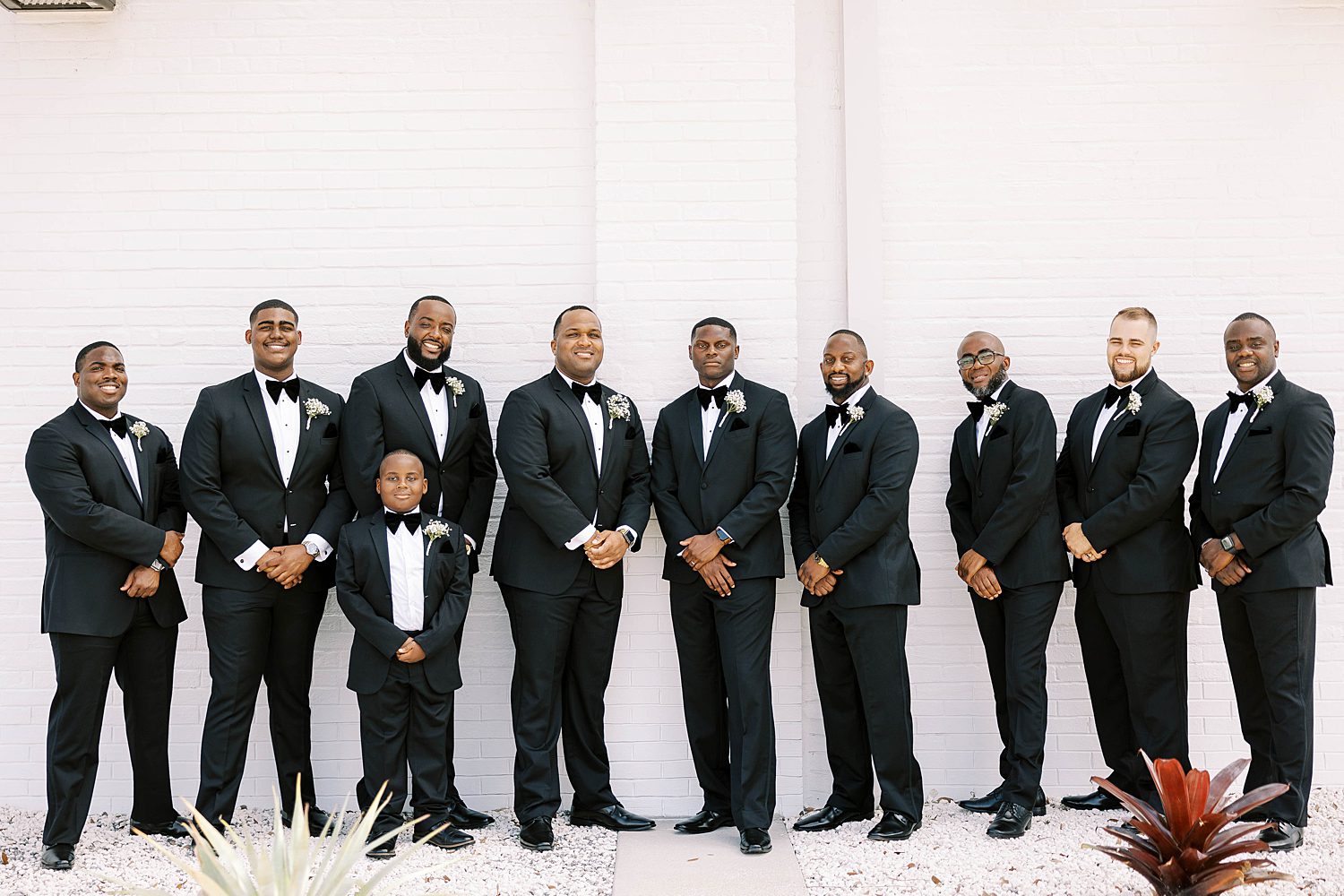 groom stands with groomsmen in classic tuxes against white wall of Haus 820
