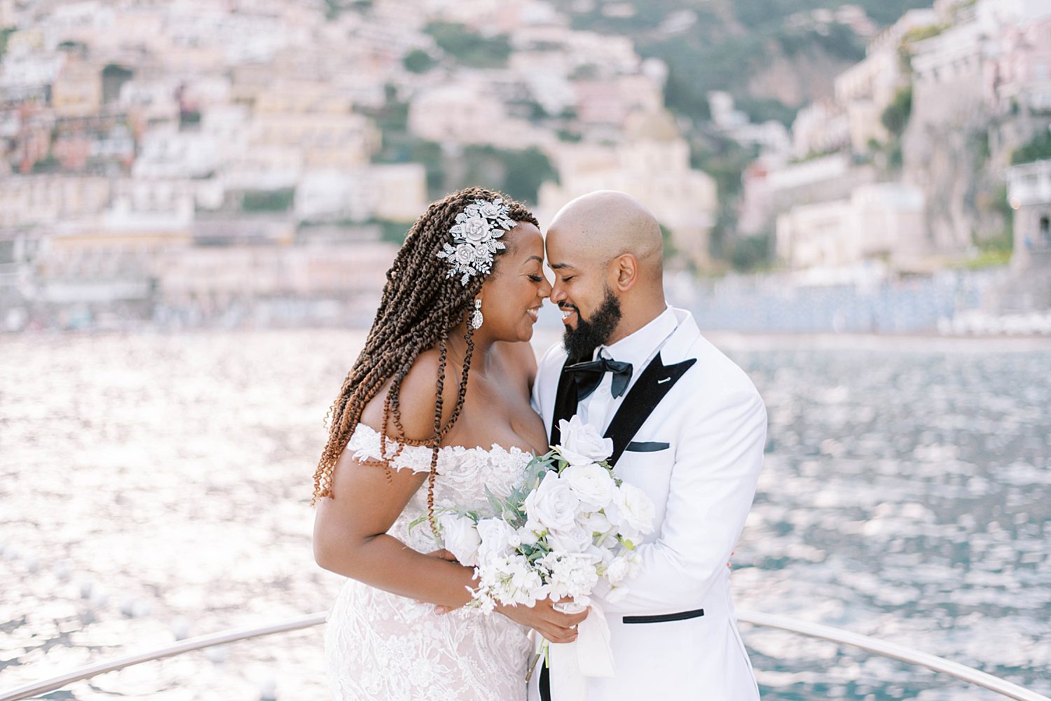 bride and groom stand on front of boat on water in front of Amalfi Coast