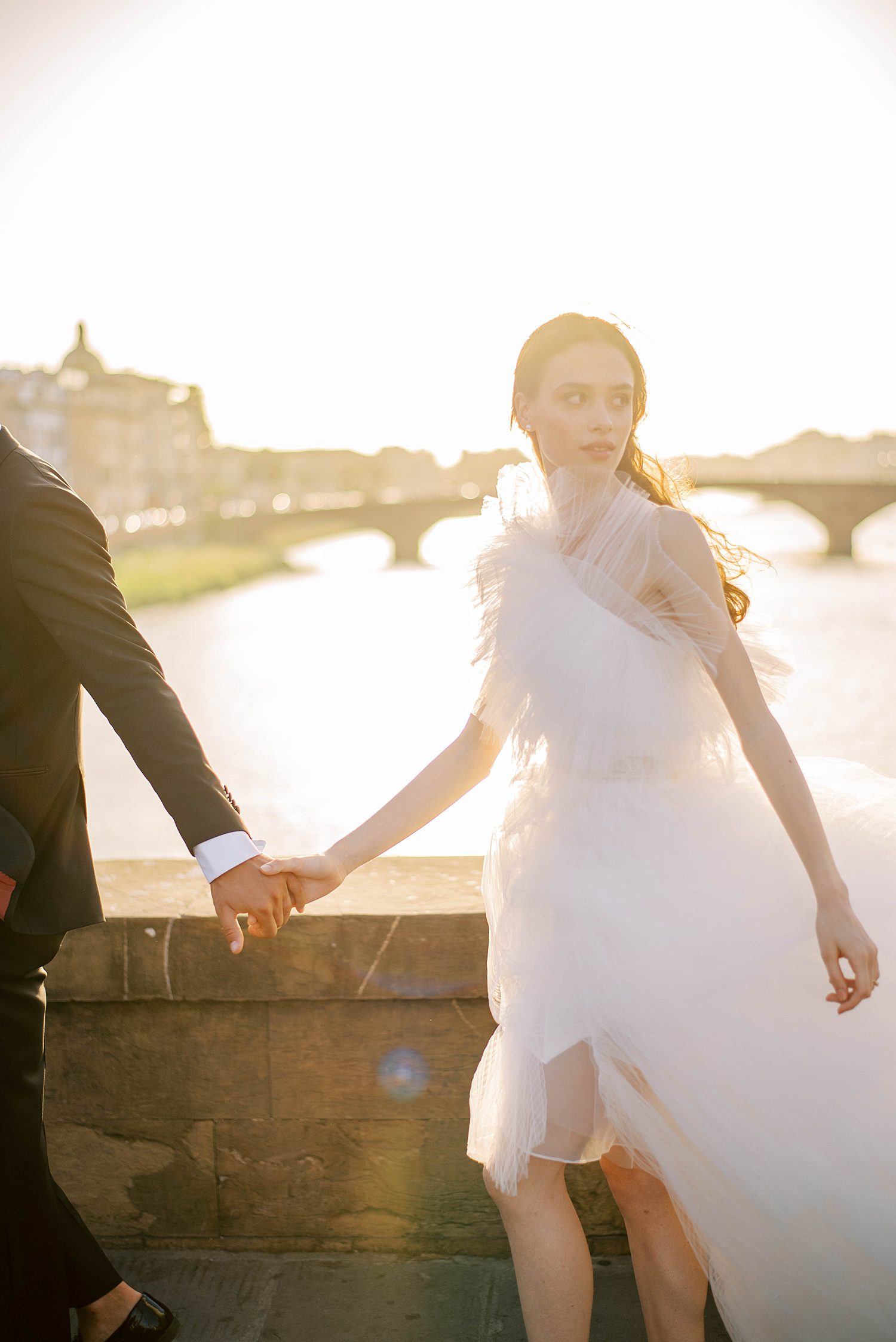newlyweds hold hands walking over stone bridge at sunset