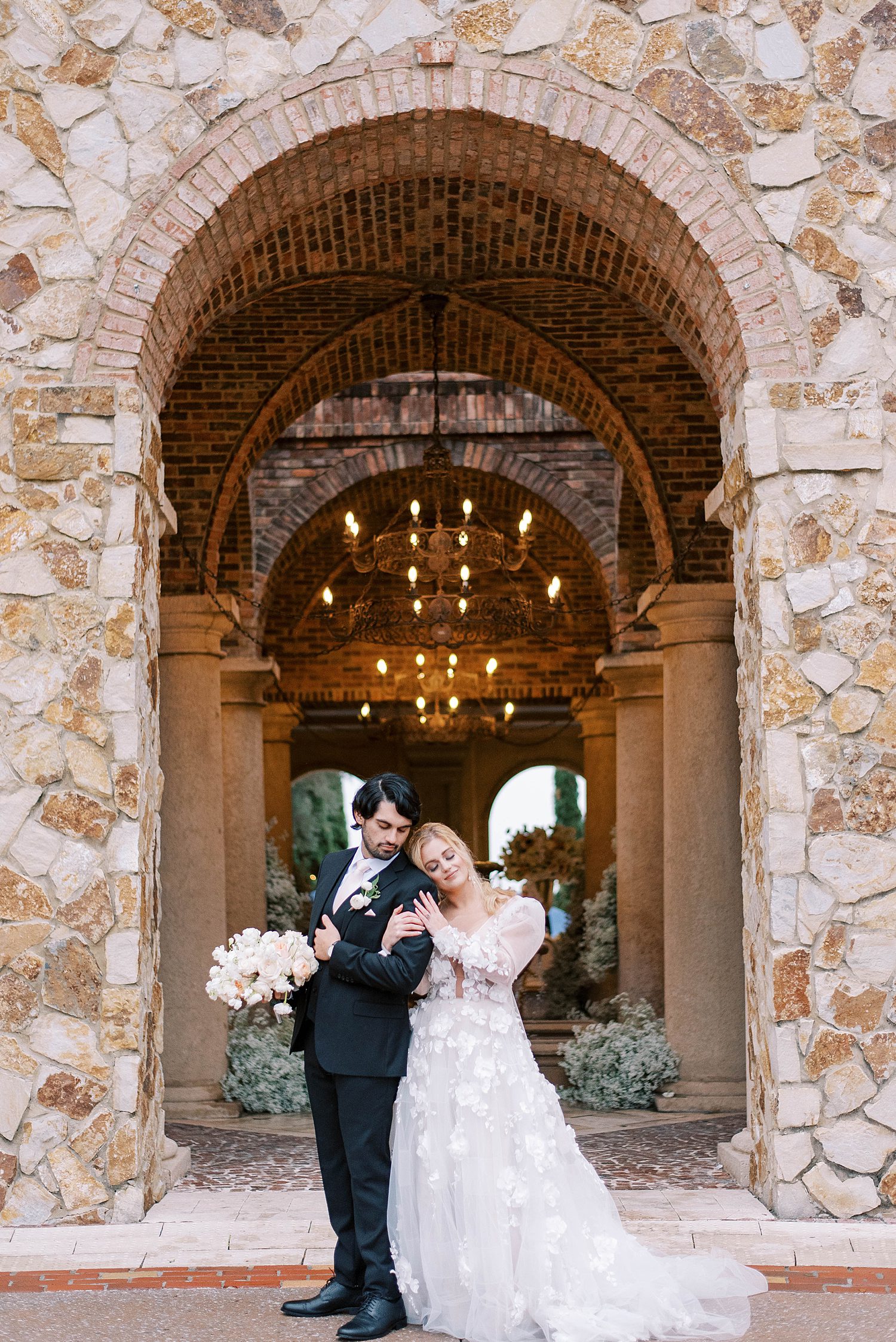 bride leans head on groom's shoulder under stone architecture at Bella Collina